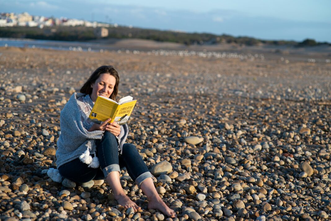 woman in blue denim jacket sitting on rocky shore reading book during daytime